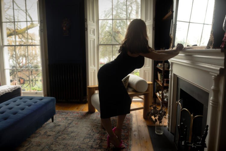 Elodie leaning against a fireplace in a black dress, silhouetted in front of a window